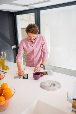 Beyaz genç gülümseyen, mutfakta ayakta, cep telefonundan hesaplama, kahvaltı yapıyor adam.