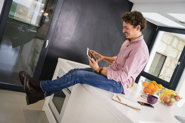 Young white man smiling, sitting on the kitchen counter, watching the news on the tablet.