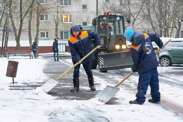 Arbeidere i uniform med store spader, en traktor fjerner snø fra veien. Snøfresing i bygatene . – stockfoto