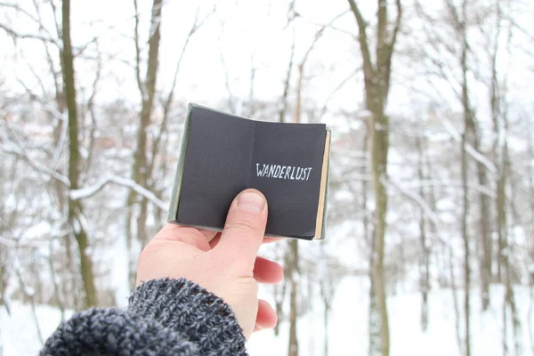 Wanderlust. Handen håller en bok med inskriptionen på bakgrunden av skogen vinter — Stockfoto