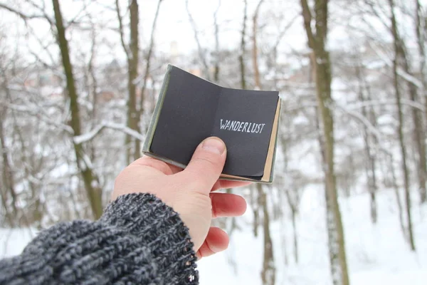 Wanderlust. Handen håller en bok med inskriptionen på bakgrunden av skogen vinter — Stockfoto