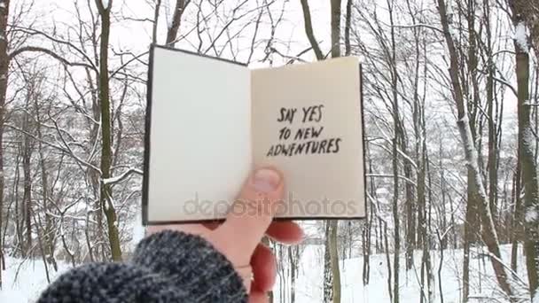 Mão segurando um livro com a inscrição Diga sim a novas aventuras no fundo da floresta de inverno — Vídeo de Stock