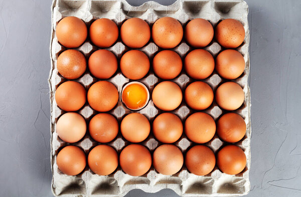 Chicken eggs in a paper tray, one egg is broken, gray background, top view