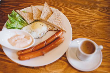 Beyaz fincanda espresso kahveli klasik İngiliz kahvaltısı, iki sosis, kızarmış yumurta, tost, ahşap arka planda marul.