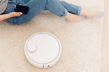 Halıda çalışan otomatik robot elektrikli süpürge kullanan genç bir kız. Akıllı ev fonksiyonlarını telefonla kontrol etmek