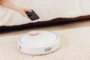 Halıda çalışan akıllı robot elektrikli süpürge kullanan genç bir kız. Telefonlu kontrol aygıtı