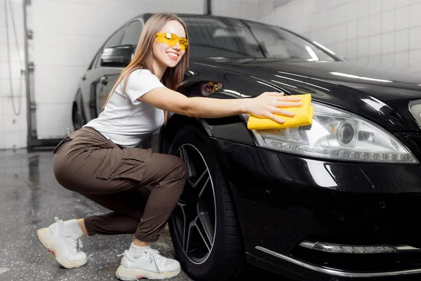 Автомойка, рабочая красивая девушка полирует и чистые микроволокна фары на черном авто — стоковое фото