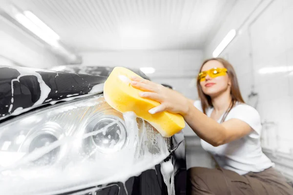 Schöne junge Frau wäscht Schaum mit gelben Schwamm Scheinwerfer des Autos — Stockfoto