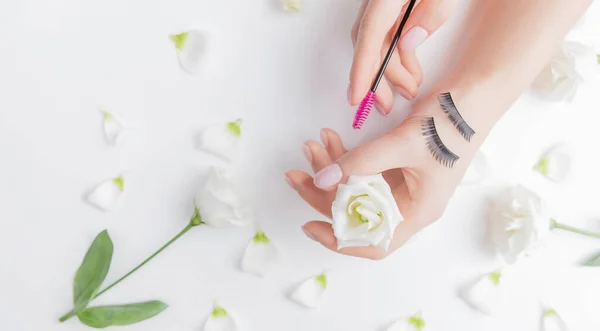 ピンクのブラシで白い花の背景に手の女の子マスターまつ毛拡張子、トップビュー。美の概念 — ストック写真