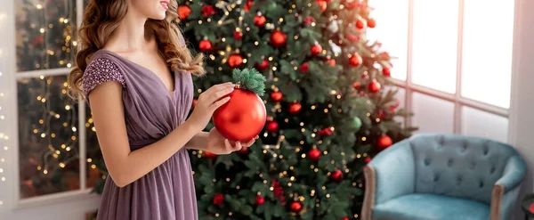 Decoração de árvore de Natal, menina bonita segurando bola vermelha em suas mãos, luz da janela. Festa conceito em vestido de noite — Fotografia de Stock