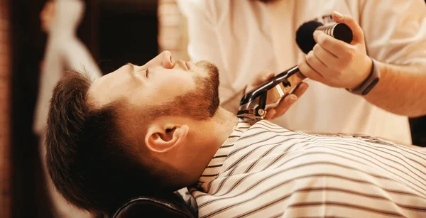 Barbier scheert zijn baard tot hippe man in de kapperszaak. Getinte foto, vintage stijl — Stockfoto