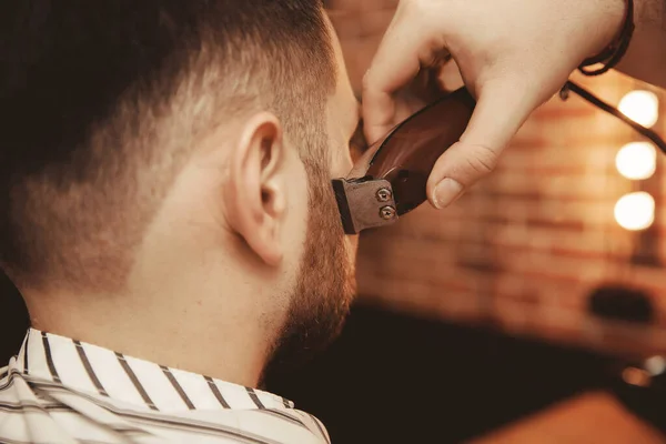 Barbier scheert zijn baard tot hippe man in de kapperszaak. Getinte foto, vintage stijl — Stockfoto