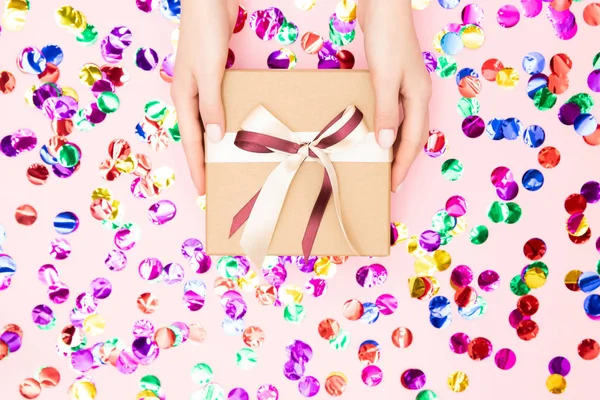 Hermosas manos niña sostienen caja de regalo presente papel artesanal sobre fondo de confeti rosa, disposición plana. Concepto Navidad, Año Nuevo — Foto de Stock