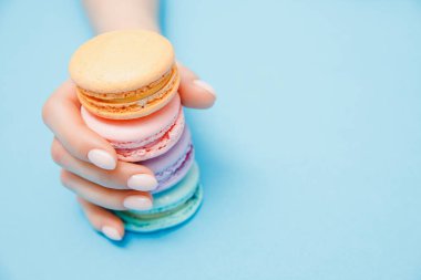 Moda manikürlü güzel el kızı mavi arka planda renkli badem kurabiyesi yiyor.