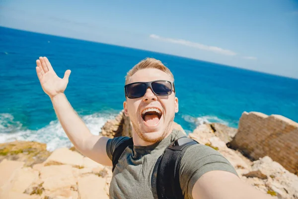 Man frilansare tar selfie foto på bakgrunden av azurblå hav, blå himmel, solglasögon och ryggsäck. Livsstilskoncept — Stockfoto