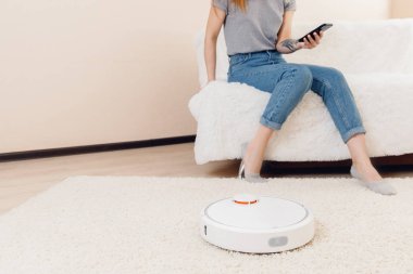 Halıda çalışan otomatik robot elektrikli süpürge kullanan genç bir kız. Akıllı ev fonksiyonlarını telefonla kontrol etmek