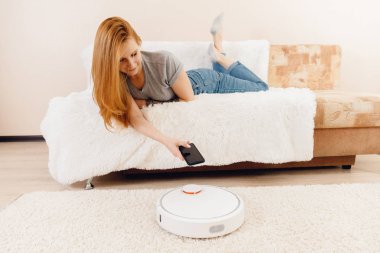 Halıda çalışan akıllı robot elektrikli süpürge kullanan genç bir kız. Telefonlu kontrol aygıtı