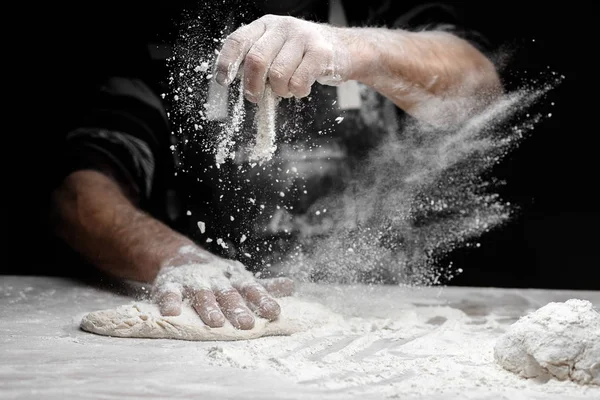 Farinha branca voa no ar no fundo preto, chefe de pastelaria bate palmas mãos e prepara massa de levedura para massas de pizza — Fotografia de Stock