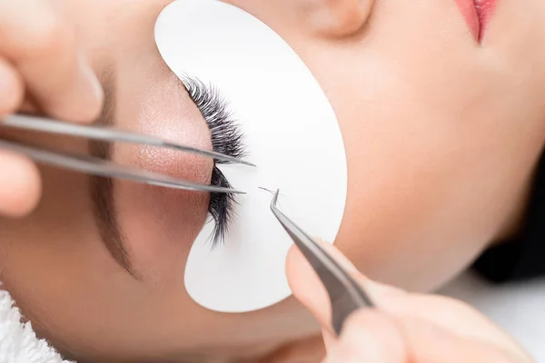 Eyelash extension procedure. Master tweezers sets fake lashes on beautiful woman