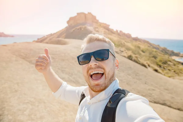 Man resenären gör selfie foto på bakgrunden av blått hav, sandstrand i solglasögon och med ryggsäck. Resekoncept. Guldbukten Malta — Stockfoto