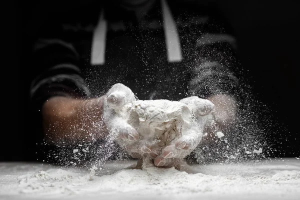 White flour flies in air on black background, pastry chef claps hands and prepares yeast dough for pizza pasta — Stock Photo, Image