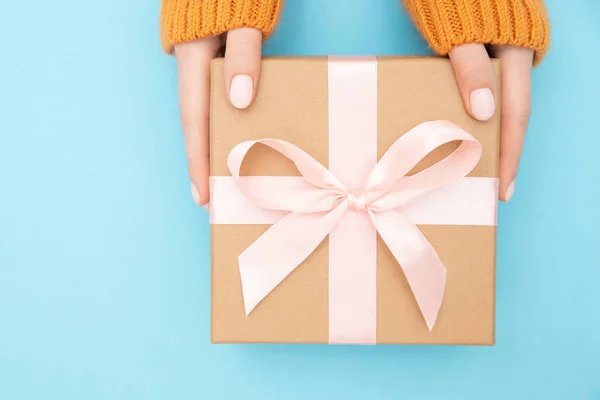 Hermosas manos niña sostienen caja de regalo presente papel artesanal con lazo rosa sobre fondo azul. Estilo plano laico — Foto de Stock