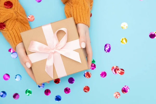 Hermosas manos niña sostienen caja de regalo presente papel artesanal con lazo rosa sobre fondo azul confeti, puesta plana. Concepto Navidad, Año Nuevo — Foto de Stock