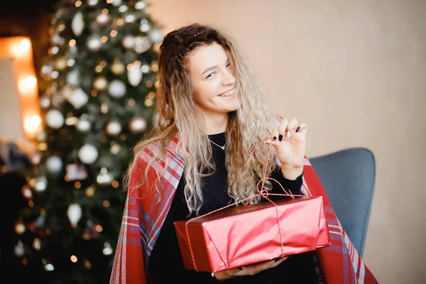 Doce jovem mulher abrir presente de ano novo, envolto em cobertor quente. Árvore de Natal no fundo borrado — Fotografia de Stock