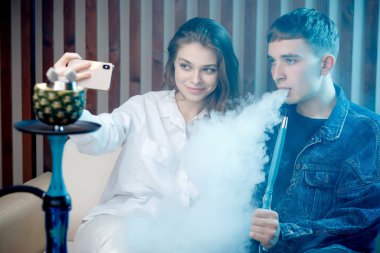 Beautiful young woman with guy hookah, blow smoke from tobacco and take selfie photo on phone, blue shisha background clipart