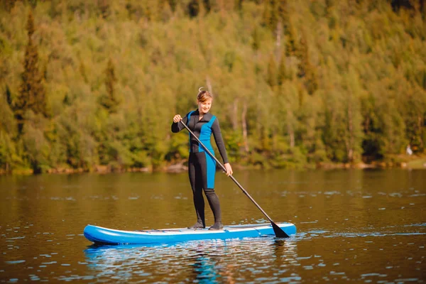 穿着保暖衣服的女孩划桨在蓝湖湖水冲浪板上的森林背景 — 图库照片