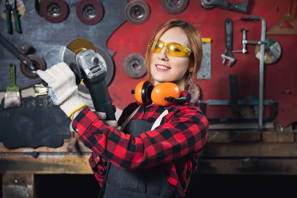 Mujer trabajadora con gafas de seguridad y auriculares utilizan molienda de rueda eléctrica en la estructura de acero en fábrica, chispa ligera — Foto de Stock