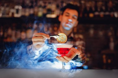 Barmen kırmızı alkollü kokteyl hazırlıyor. Martini bardağında mavi duman ve portakalla süslenmiş.