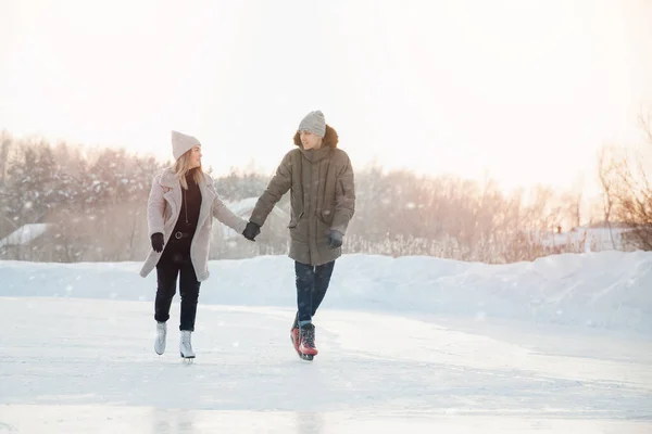 Menina e homem estão de mãos dadas olhando um para o outro e comendo no gelo congelado do lago em patins. No fundo é pôr do sol — Fotografia de Stock