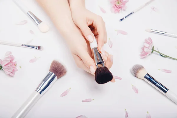 Las manos de las niñas sostienen pinceles de maquillaje en el fondo de flores de color rosa, concepto de salón de belleza. Vista superior —  Fotos de Stock