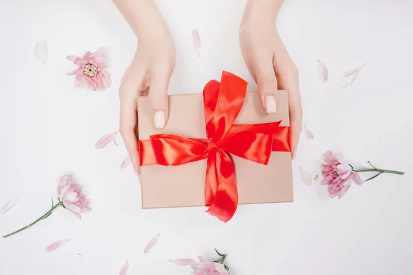 Manos de niña sosteniendo regalo regalo caja de artesanía con lazo rojo sobre fondo blanco con flores, disposición plana. Concepto día de la madre, día de San Valentín — Foto de Stock