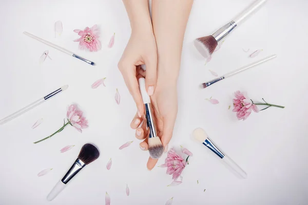 Las manos de las niñas sostienen pinceles de maquillaje en el fondo de flores de color rosa, concepto de salón de belleza —  Fotos de Stock