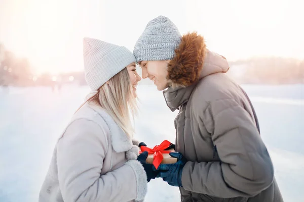 Os amantes dão caixa de presentes com arco vermelho uns aos outros fora do parque de inverno, vestidos com roupas quentes e chapéus de malha. Conceito Dia dos Namorados ou Natal — Fotografia de Stock