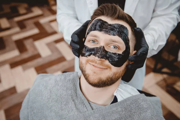 Spa and skin care for men. Barber professional applies black charcoal mask to client man