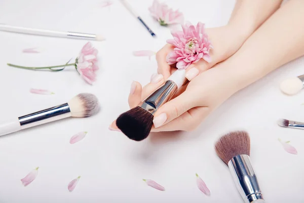 Las manos de las niñas sostienen pinceles de maquillaje en el fondo de flores de color rosa, concepto de salón de belleza —  Fotos de Stock