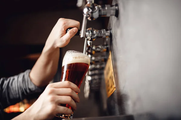 Il barista versa la birra dal rubinetto al bicchiere, sfondo scuro. Alcol mestiere bevanda concetto — Foto Stock