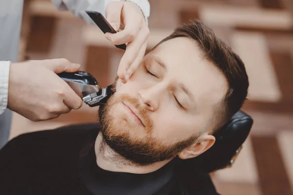 Barbería esquilando barba a hombre en barbería enmarcando la línea del cabello. maquinilla de afeitar eléctrica, vintage teñido marrón — Foto de Stock