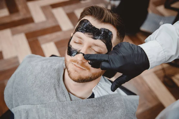Peluquería aplica mascarilla de carbón negro a la cara del hombre para limpiar la piel de los poros y eliminar el acné de la nariz, salón de spa — Foto de Stock