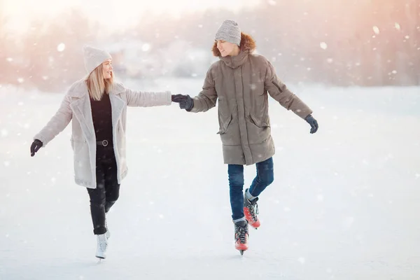 Loving couple in winter with skates on rink on sunset background, guy and girl dressed in warm hat