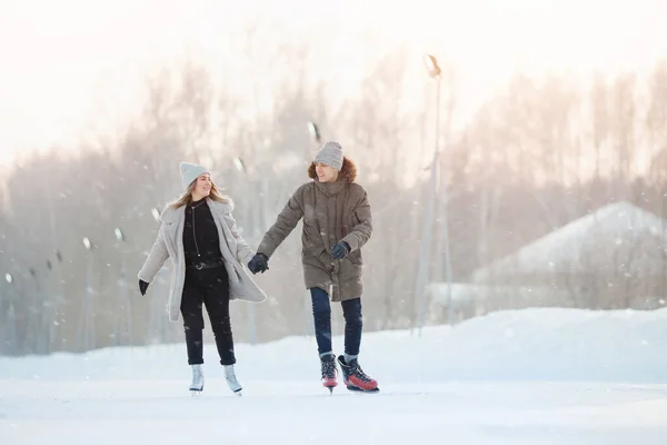 Glada unga kärlekspar håller varandra i handen, åker skridskor på rinken, det snöar på vintern. Begreppet semester utomhus aktiviteter — Stockfoto