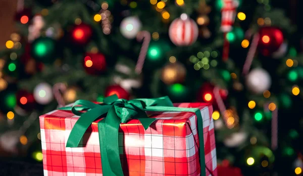 Νέο Έτος δώρο με κόκκινο πλέγμα γραμμές, δεμένα πράσινη κορδέλα στο φόντο του χριστουγεννιάτικου δέντρου με φωτισμό — Φωτογραφία Αρχείου