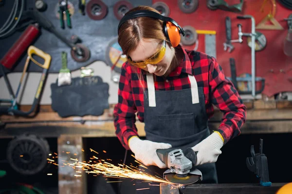 Mujer trabajadora con gafas de seguridad y auriculares utilizan molienda de rueda eléctrica en la estructura de acero en fábrica, chispa ligera — Foto de Stock