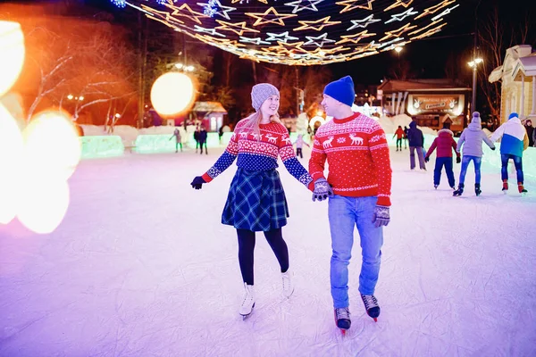 Beautiful young couple of lovers in warm red sweater on skating rink figured white skates night on background illuminations winter — Stock Photo, Image