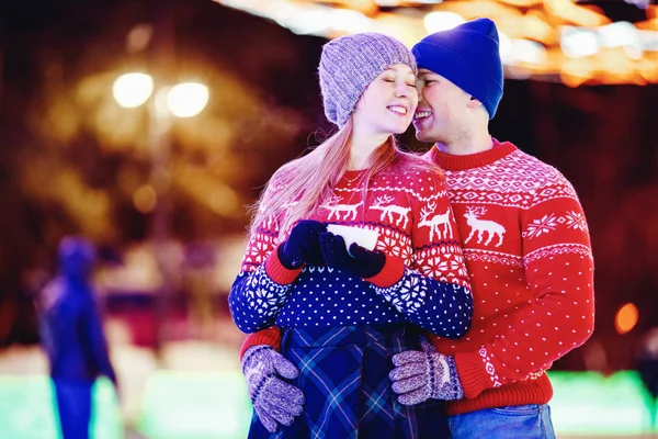 Loving couple drinks hot tea or coffee from thermos mugs on winter street in ice rink night illumination — Stock Photo, Image