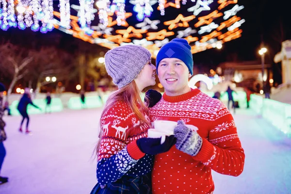 Loving couple drinks hot tea or coffee from thermos mugs on winter street in ice rink night illumination — Stock Photo, Image