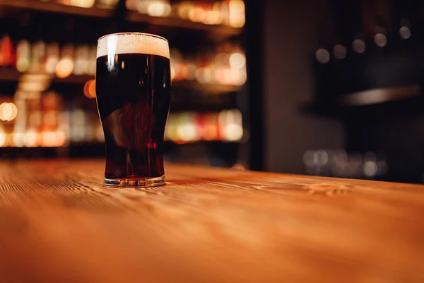 Bicchiere di birra scura artigianale con schiuma sul bancone del bar in legno, pub sfondo sfocato — Foto Stock